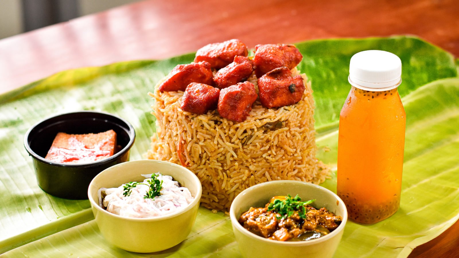 Chicken 65 biriyani on a banana leaf with onion raita, brinjal curry, a sweet and a bottle of nannari sharbat